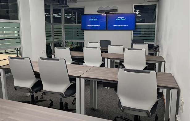 Sala de formación con varias mesas individuales y sillas de oficina de color blanco y negro. Al fondo, dos pantallas de televisión muestran el logo "West Telco" y el eslogan "La tecnología como puente". Hay grandes ventanales con persianas que dividen la sala de otras áreas.