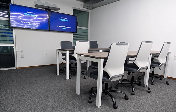 Sala de reuniones con dos mesas de trabajo cuadradas, colocadas una al lado de la otra. Las mesas tienen tableros de madera clara y patas de metal blanco. Alrededor hay seis sillas de oficina con ruedas, de color blanco y negro. En la pared, dos pantallas de televisión muestran el logo "West Telco". El suelo está cubierto con una moqueta gris.