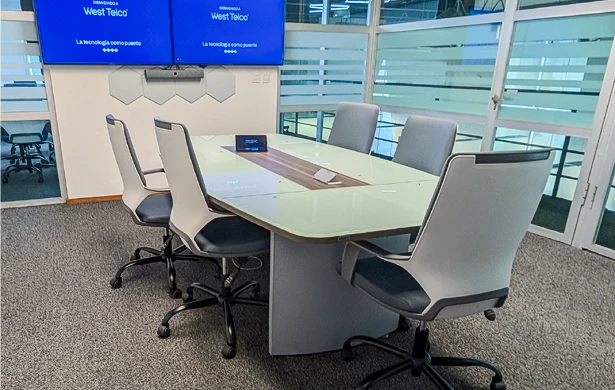 Mesa de juntas de una oficina moderna. Alrededor de la mesa hay seis sillas con ruedas, de color blanco y negro. La mesa es de madera clara con una superficie de vidrio, que tiene una sección de madera oscura en el centro. Al fondo, en una pared blanca, hay dos pantallas de televisión con el logo "West Telco" y el eslogan "La tecnología como puente". El suelo está cubierto de moqueta gris.