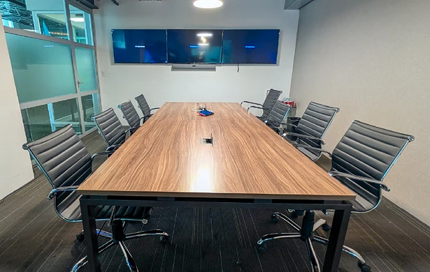 Mesa de juntas de madera oscura en una sala de reuniones. Alrededor de la mesa hay sillas de oficina de cuero negro. Tres pantallas de televisión oscuras cuelgan en la pared del fondo.