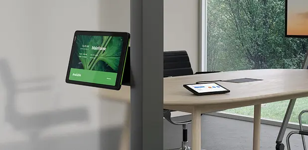 <br />
Una tableta de control montada en la pared muestra la disponibilidad de una sala de reuniones con una interfaz de usuario verde. En el fondo, se ve parcialmente una mesa de conferencias de madera clara con otra tableta sobre ella, y una ventana con vistas a un bosque.